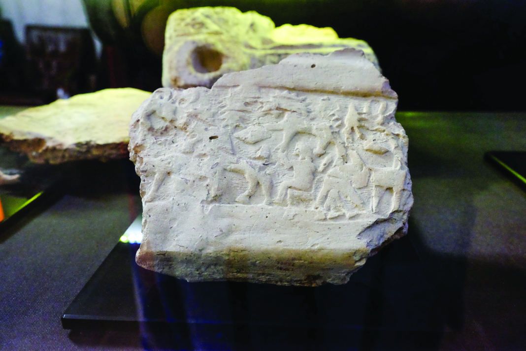 Fragment of mudbrick with carved relief figures showing human and animal forms, displayed in a museum setting