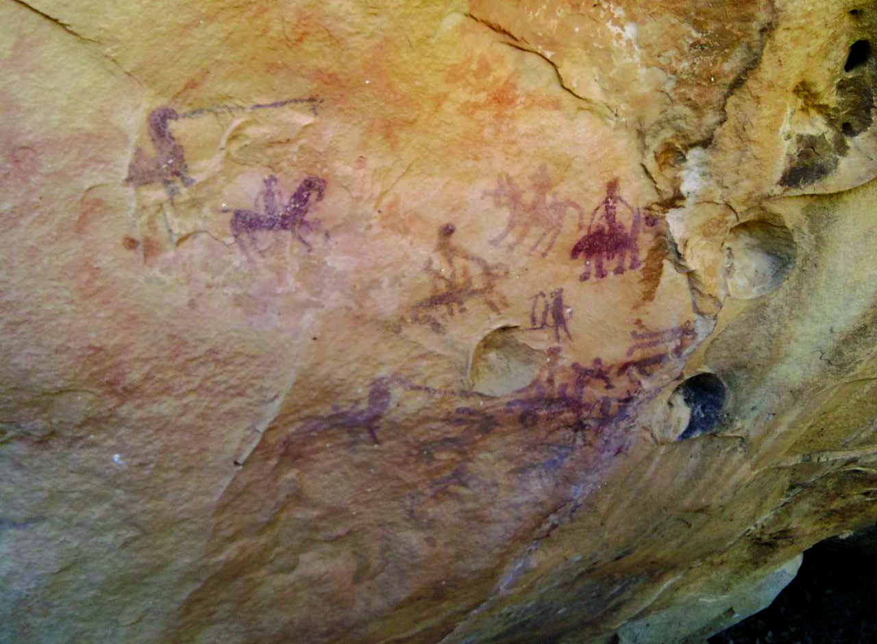 Close-up of the painted scene inside a Lorestan rock shelter showing riders, animals, and hunting figures in dark red ochre on yellow sandstone