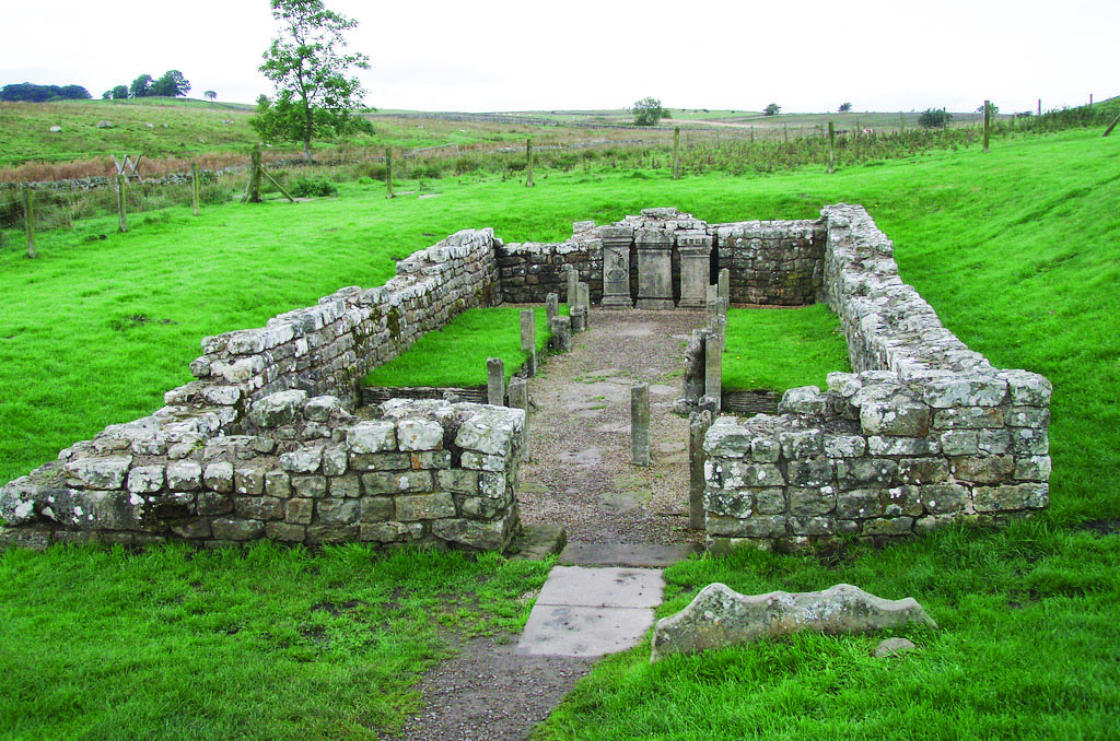 Ruins of a Mithraeum (Mithraic temple) in the English countryside, showing low stone walls in a rectangular plan amid green fields