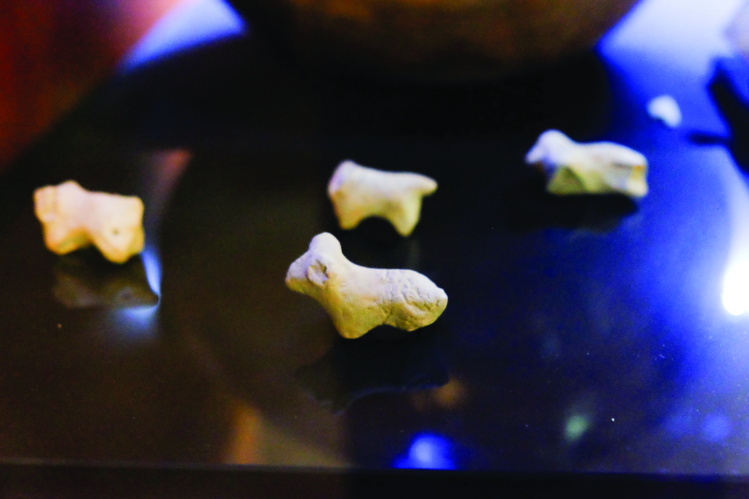 Four small clay goat figurines displayed on a reflective dark surface in a museum case, simple abstracted forms from the ancient Sialk settlement