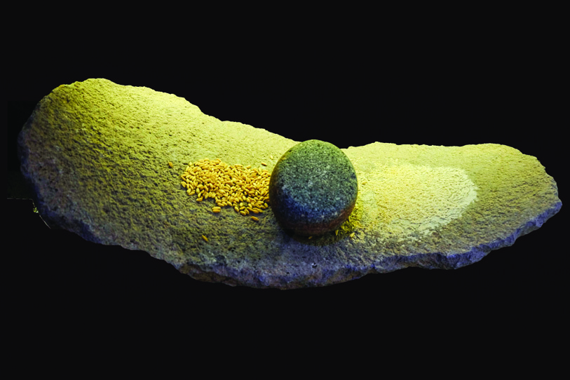 An ancient stone hand mill (quern) with a round grinding stone and scattered grain, displayed against a black background
