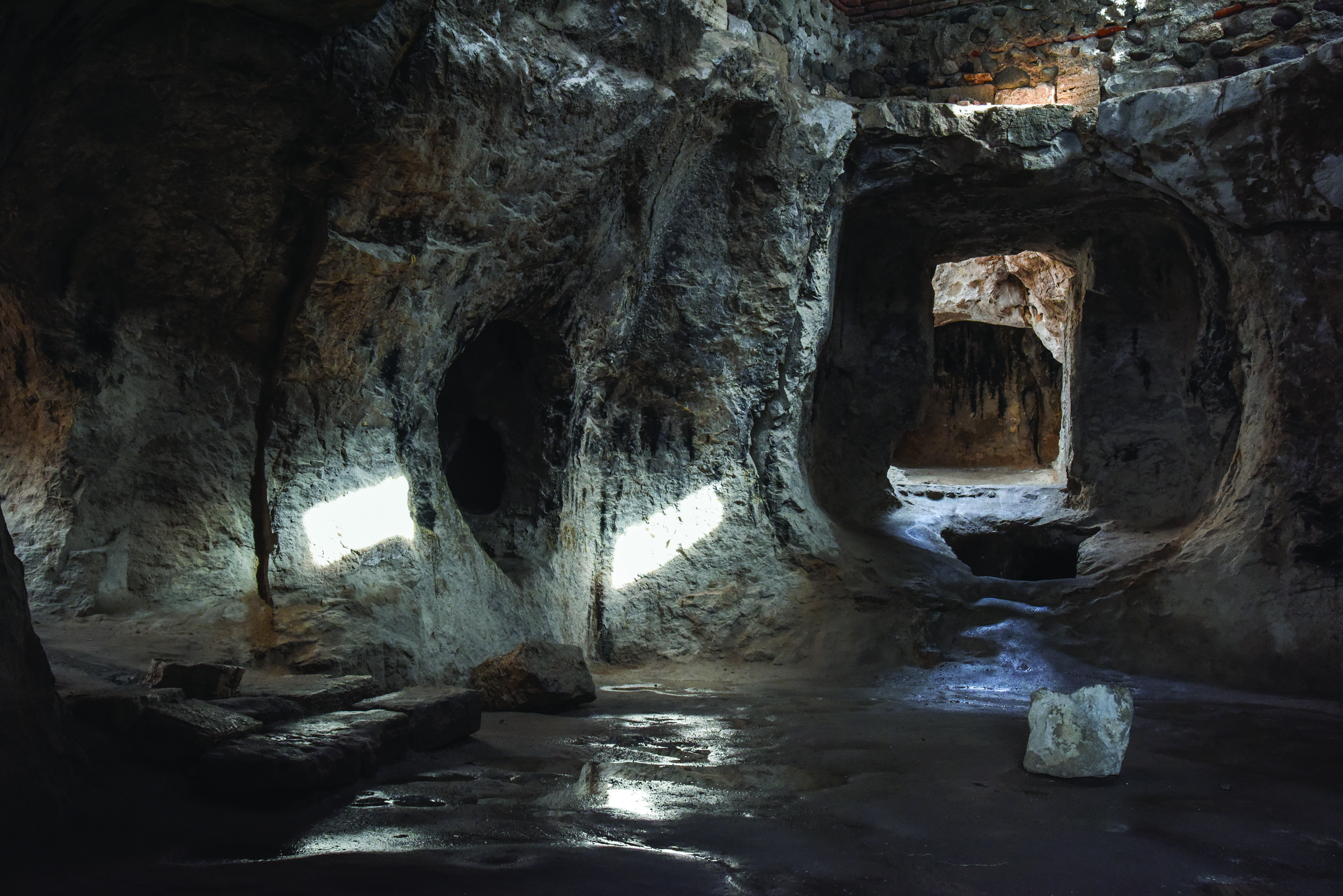 Interior of the Varjovi Mithraeum central hall showing dramatic patches of light falling on the rock walls through collapsed ceiling openings, with water on the floor reflecting the light