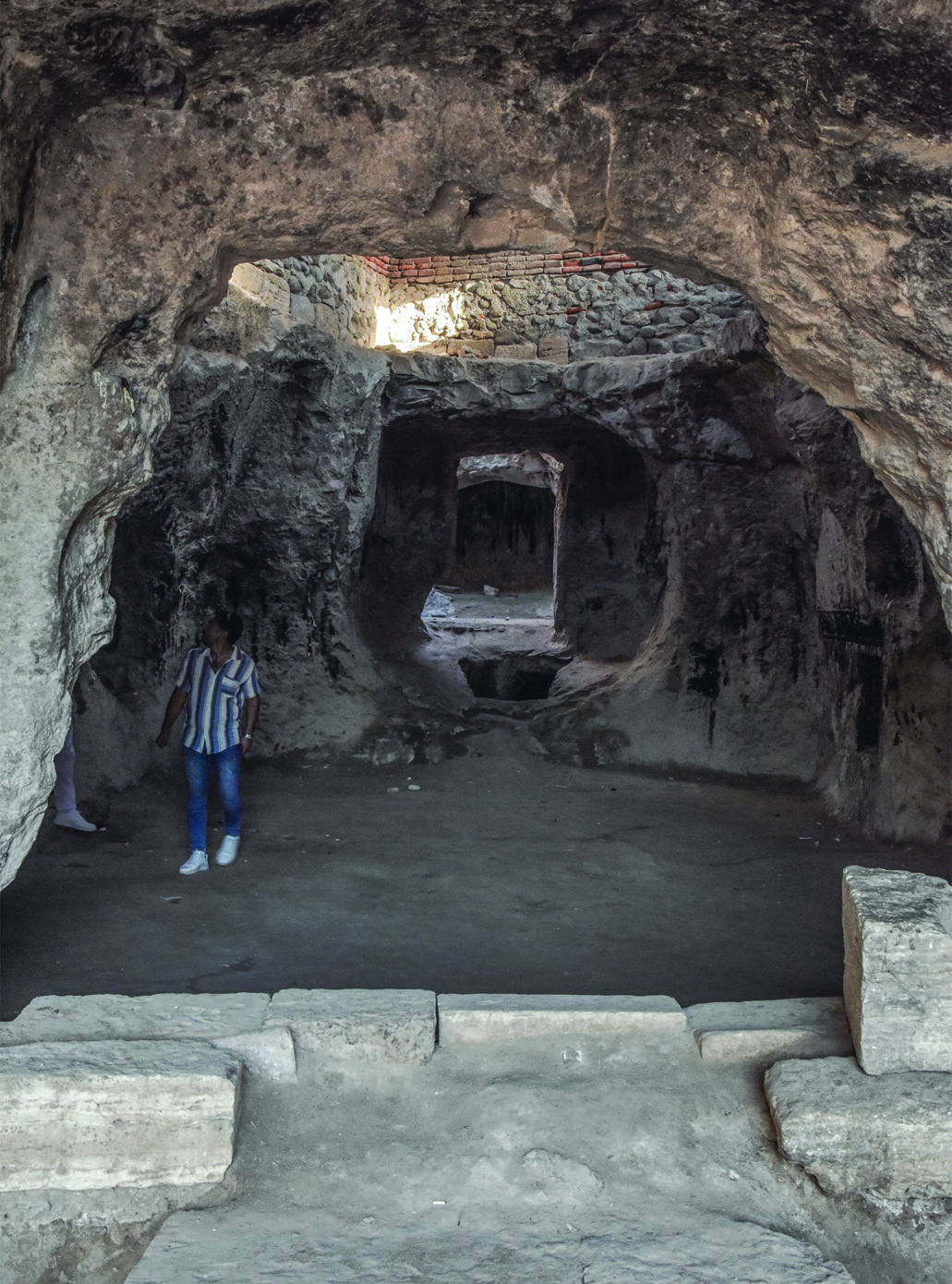 A visitor stands inside the central hall of the Varjovi Mithraeum, providing scale to the large rock-cut space with its barrel vault and stone benches