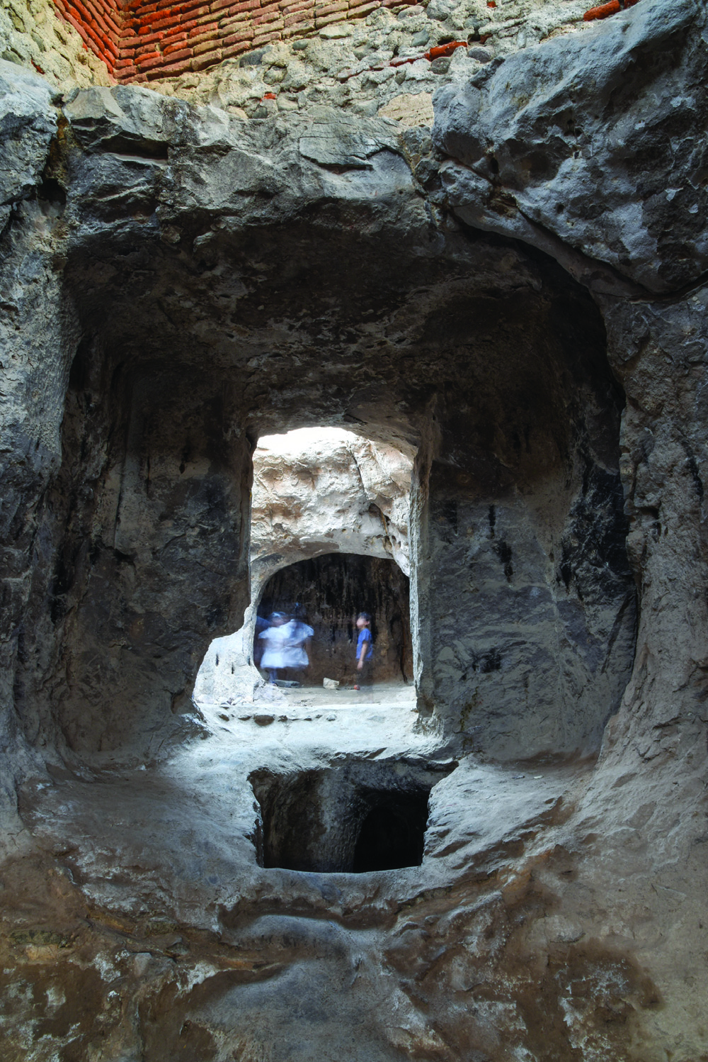 Interior view looking through a rock-cut doorway into the central hall of the Varjovi Mithraeum, with the sanctum visible beyond and a pit in the foreground