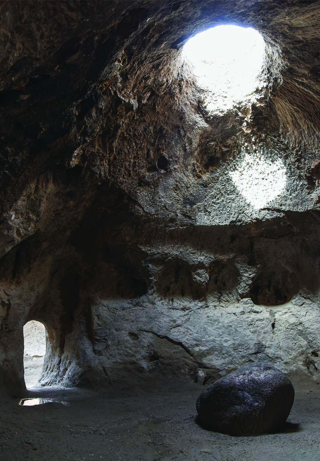 Interior of the Varjovi Mithraeum with light streaming through a ceiling oculus, illuminating the rock walls, with a doorway visible on the left and a large stone on the floor