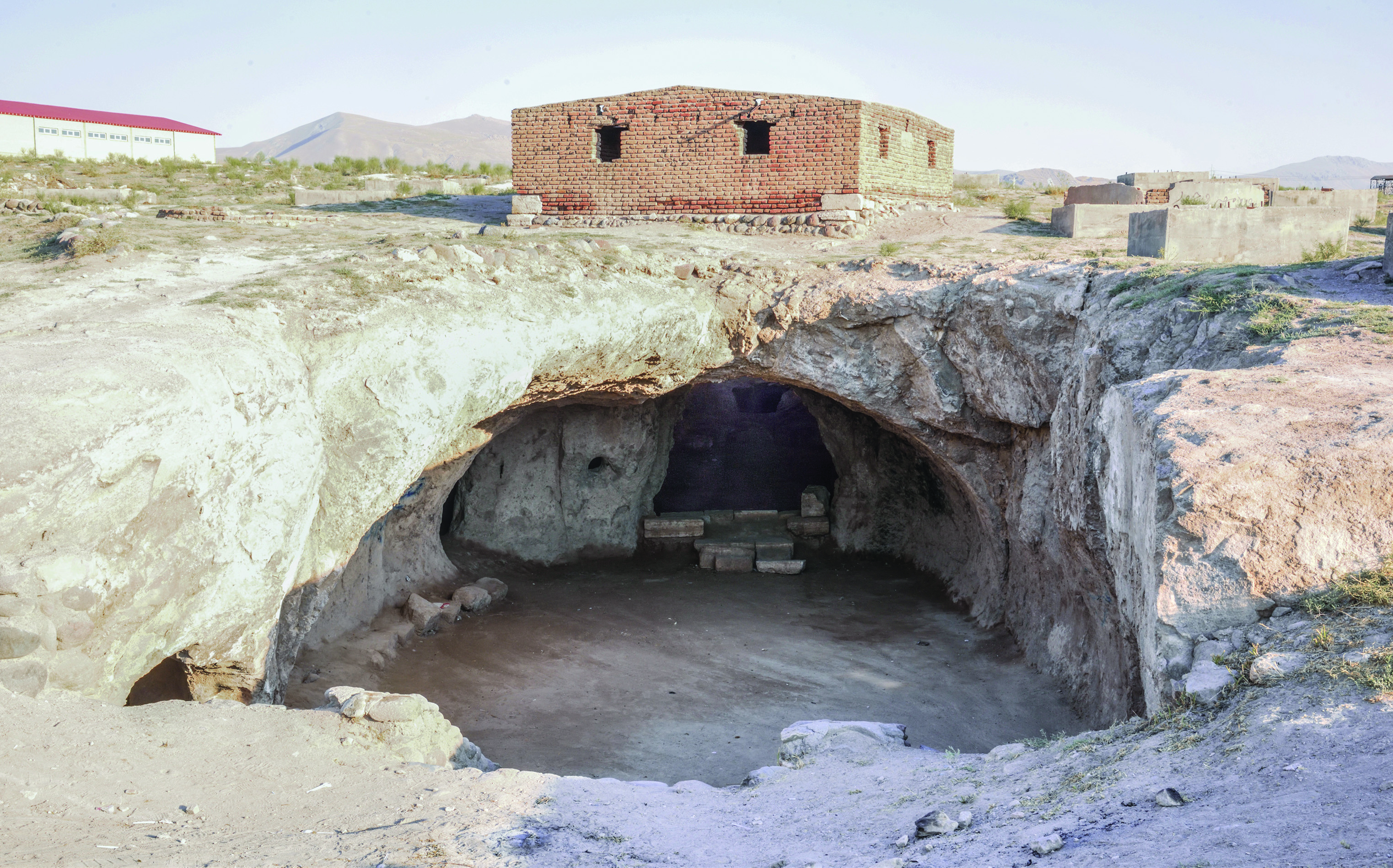 Exterior view of the Varjovi Mithraeum showing the underground temple exposed from above, with a modern brick structure built over the central hall and mountains in the background