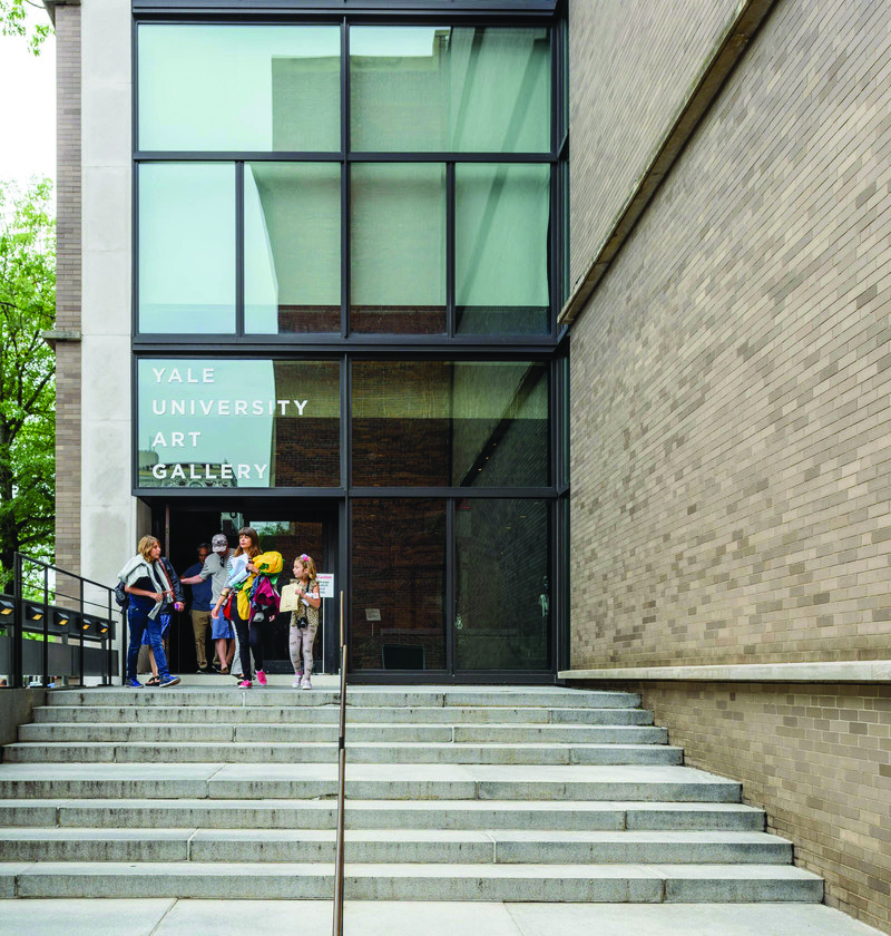 Color photograph of the Yale Art Gallery entrance exterior showing visitors entering through the glass doors beneath the brick facade and concrete frame