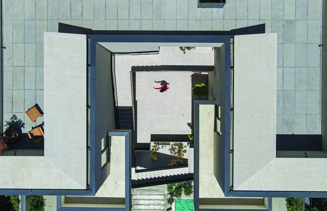 Looking straight down into the central stair void &mdash; a figure in red visible at the bottom, white tile surfaces, dark steel railings, greenery at the edges