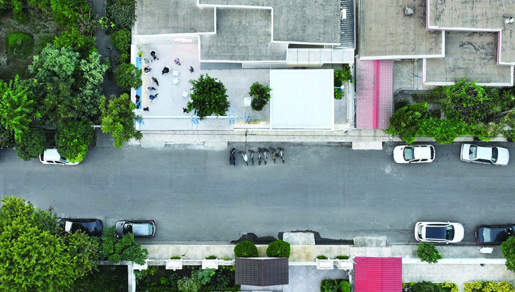 Aerial drone view of Caf&eacute; Zest showing the white rooftops, gravel courtyard, trees, blue scaffolding elements, and the tree-lined residential street with parked cars