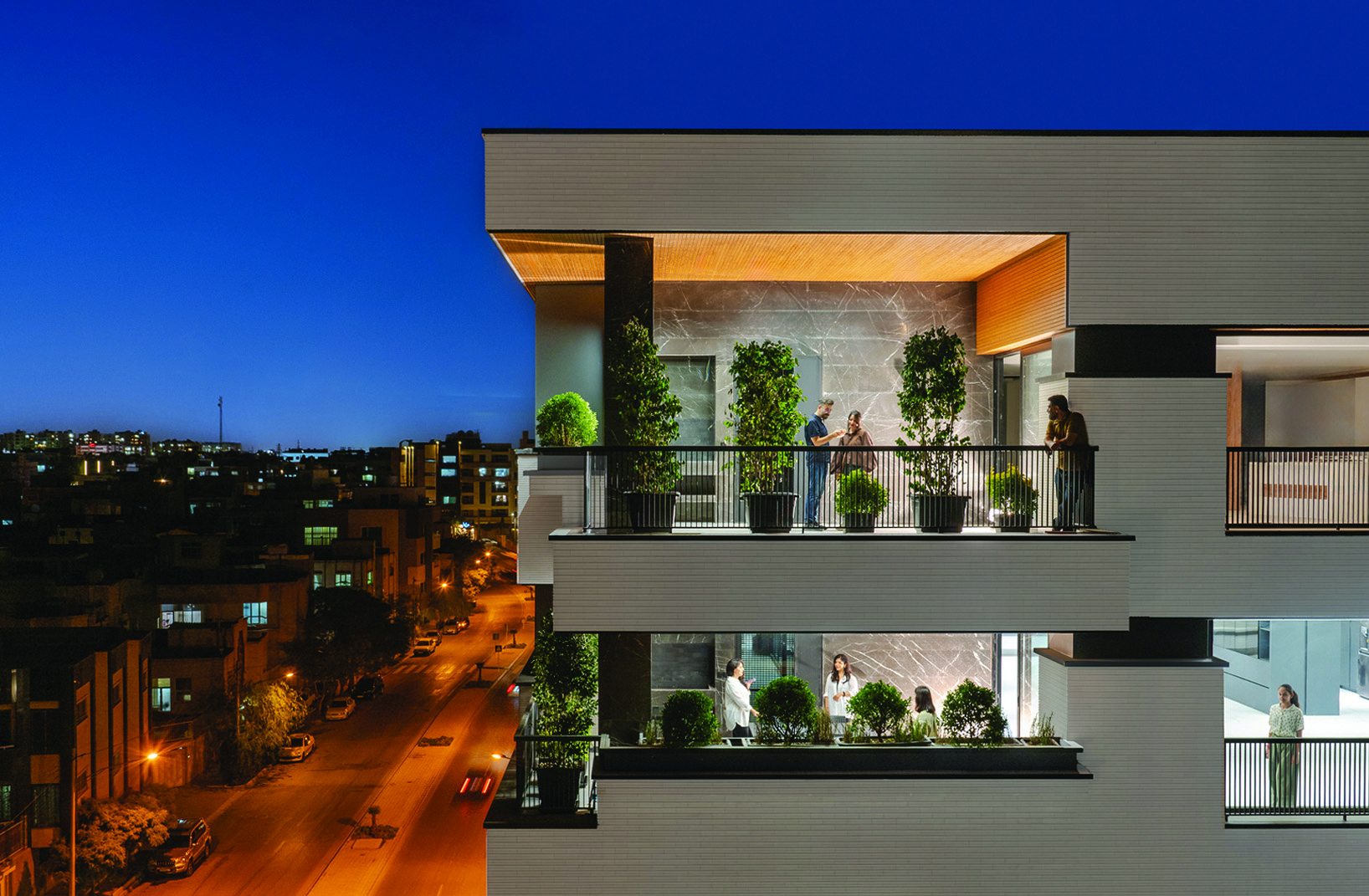 Upper floors of Bagh Mashhad at blue hour, people gathering on two levels of balconies with potted plants, the illuminated street and city skyline visible below
