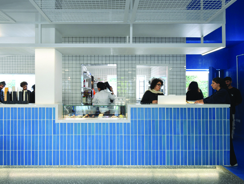 Interior view of the main bar counter clad in vertical blue tiles, white grid-tile walls, metal mesh ceiling panels, staff working behind the counter, customers seated at the bar