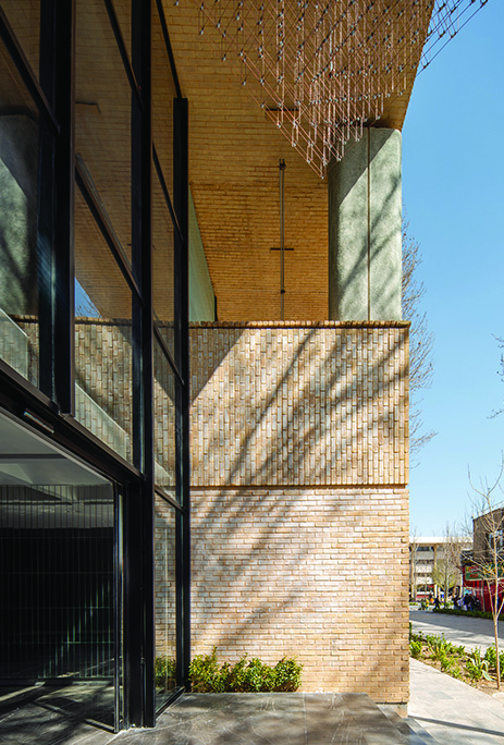 Brick facade detail at ground level with vertical brick pattern, glass curtain wall, and planted beds