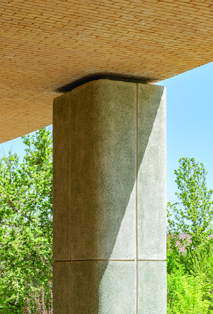 Close-up of a concrete column meeting the brick-clad soffit &mdash; curved transition between column and ceiling, greenery visible beyond