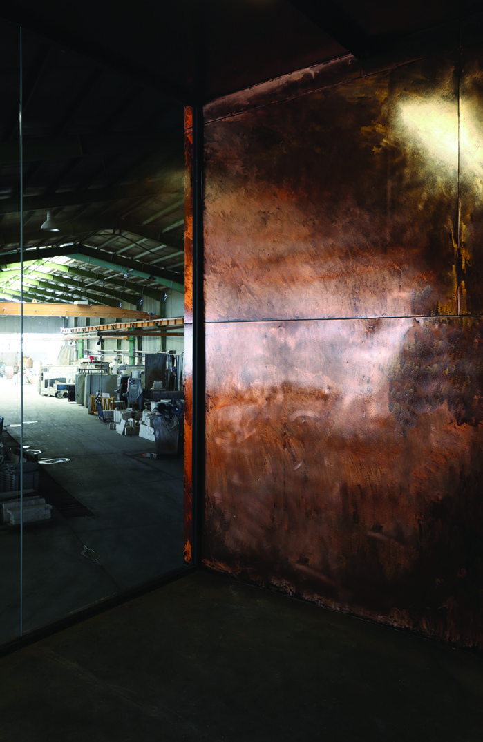Interior of a copper-clad module looking outward through full-height glass to the factory shed beyond, copper walls reflecting light