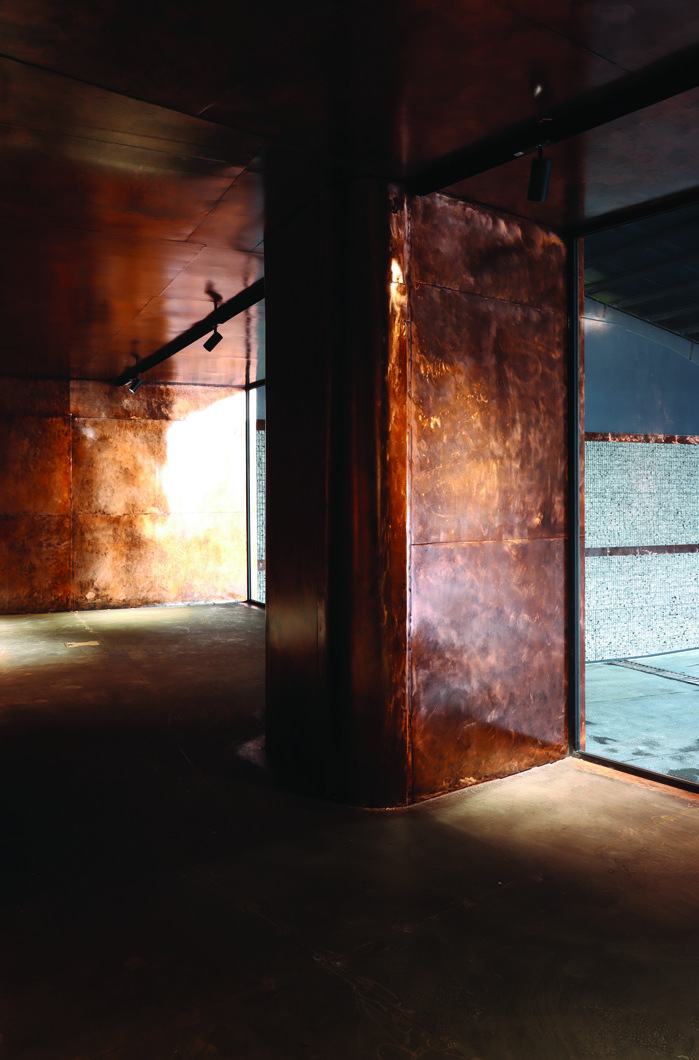 Interior of a copper-clad module &mdash; warm patinated copper walls and ceiling surrounding a steel column, glass wall looking out to the gabion surface