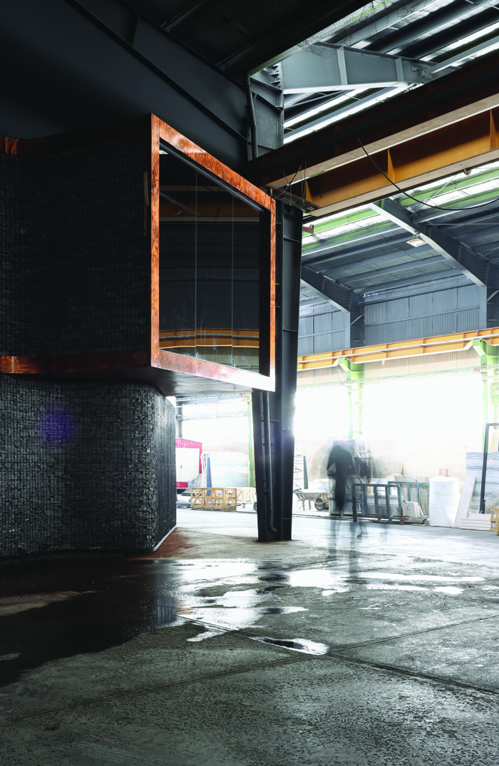 Cantilevered copper-and-glass module seen from a distance inside the factory shed, a blurred worker passing beneath it