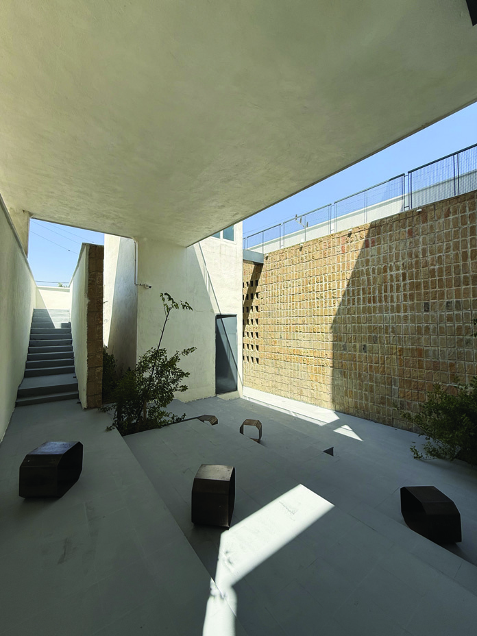 Wide view of the courtyard from beneath the concrete canopy, with the large stone wall catching dramatic sunlight, an exterior staircase visible on the left, metal stools scattered across the floor, and trees growing in raised planters