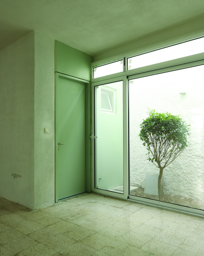 Interior corner showing a green door beside a full-height glass wall looking onto a small white courtyard with a single tree