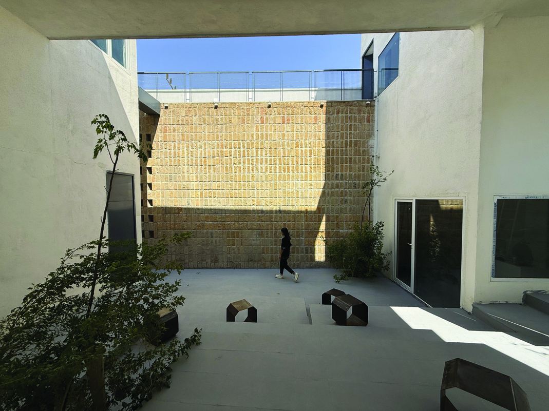 The main courtyard of Voocho seen from under a concrete canopy, with a large hewn stone wall as backdrop, a person walking across the space, angular metal stools scattered on the concrete floor, a young tree, and blue sky above