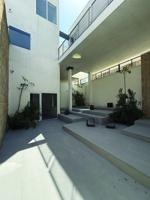 View of the courtyard from the upper level, showing stepped concrete platforms, plants growing in built-in planters, the stone wall below, glass doors to interior spaces, and the white volumes above