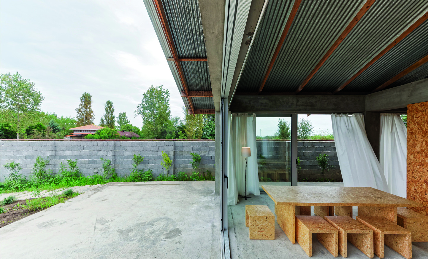 Covered terrace at Pati Project showing the double-skin corrugated metal roof extending over the outdoor area, OSB dining table visible through the open glass walls, cinder-block perimeter wall and greenery beyond