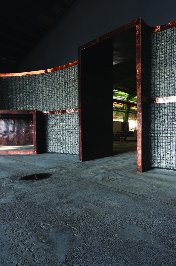 Curved gabion wall enclosing a dark doorway with copper frame, looking through to the factory floor beyond