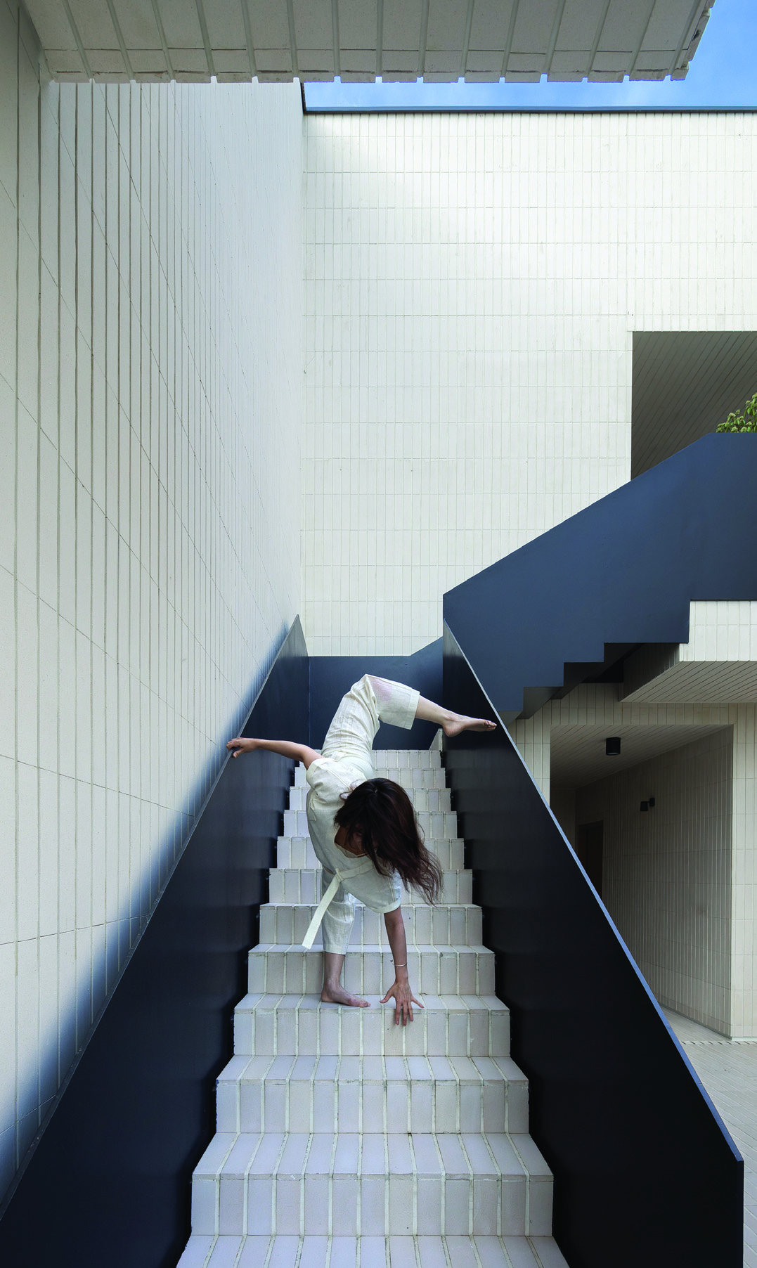 A dancer in white performing on the dark steel staircase, arching backwards with arms extended, white tile walls rising on either side, open sky above