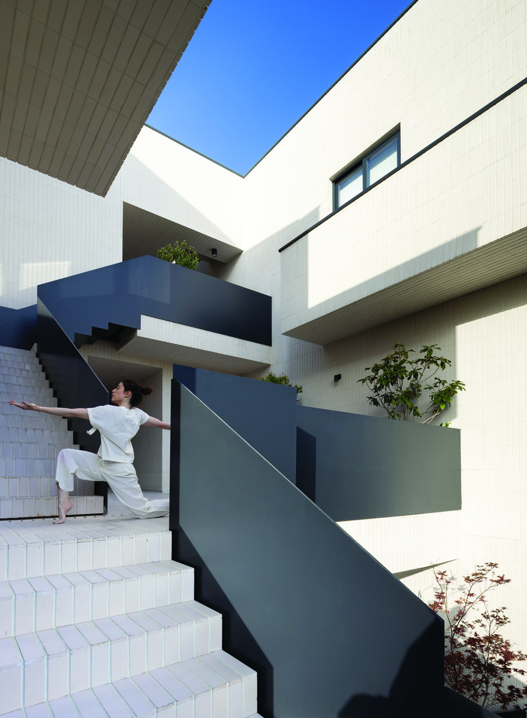 A dancer in white leaning dynamically on the dark steel staircase, with the stair zigzagging upwards between white tile volumes, planters with greenery, blue sky above