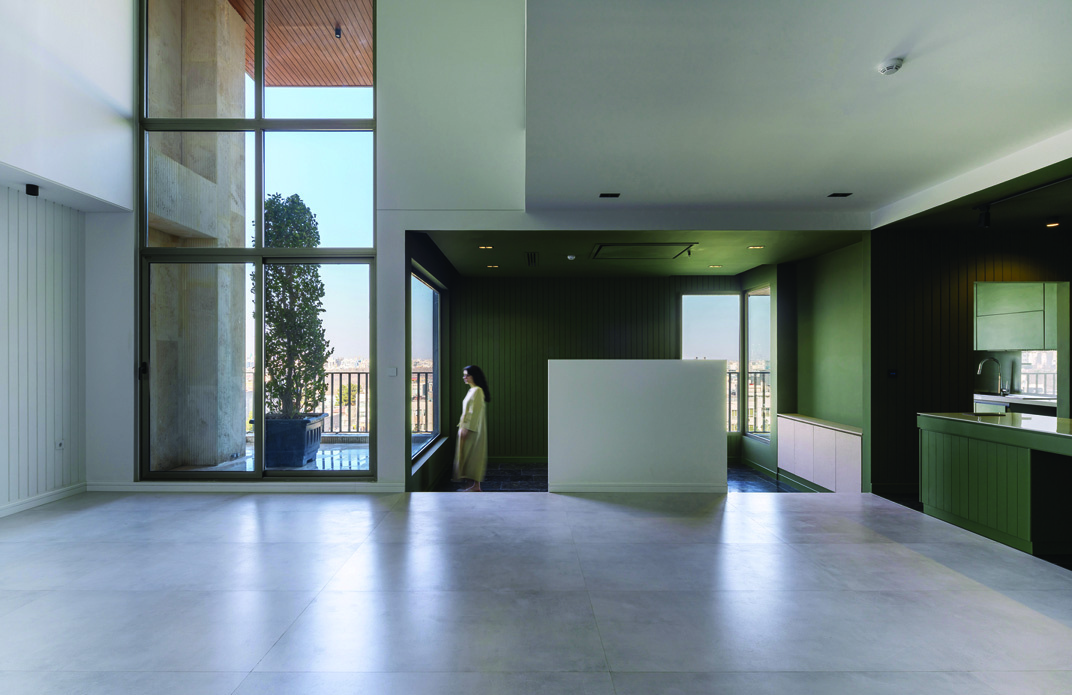 Double-height interior space with floor-to-ceiling windows opening onto a terrace, green panelled kitchen zone beyond a white counter, a woman standing near the glass