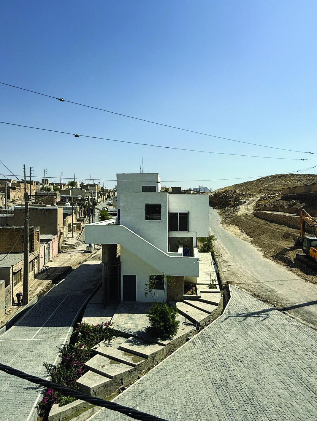 Elevated view of Voocho from above, showing the white stepped volume with an exterior staircase, the dense neighbourhood to the left and a bare hillside to the right, construction equipment visible