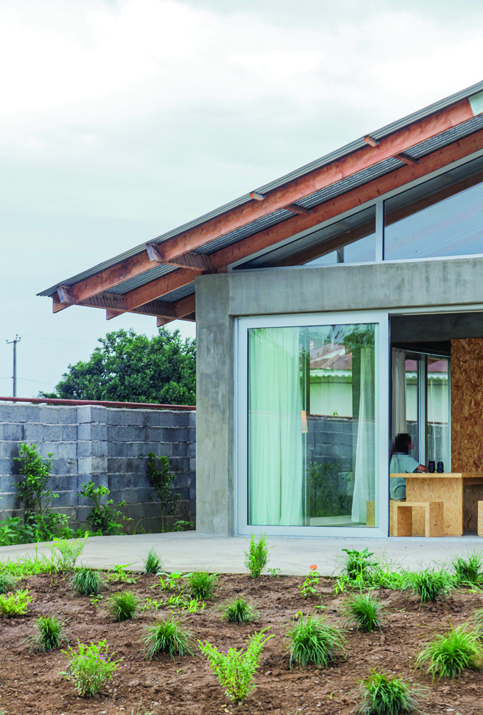 Close-up of the glass wall and concrete column with the corrugated metal roof extending overhead, newly planted garden bed in the foreground