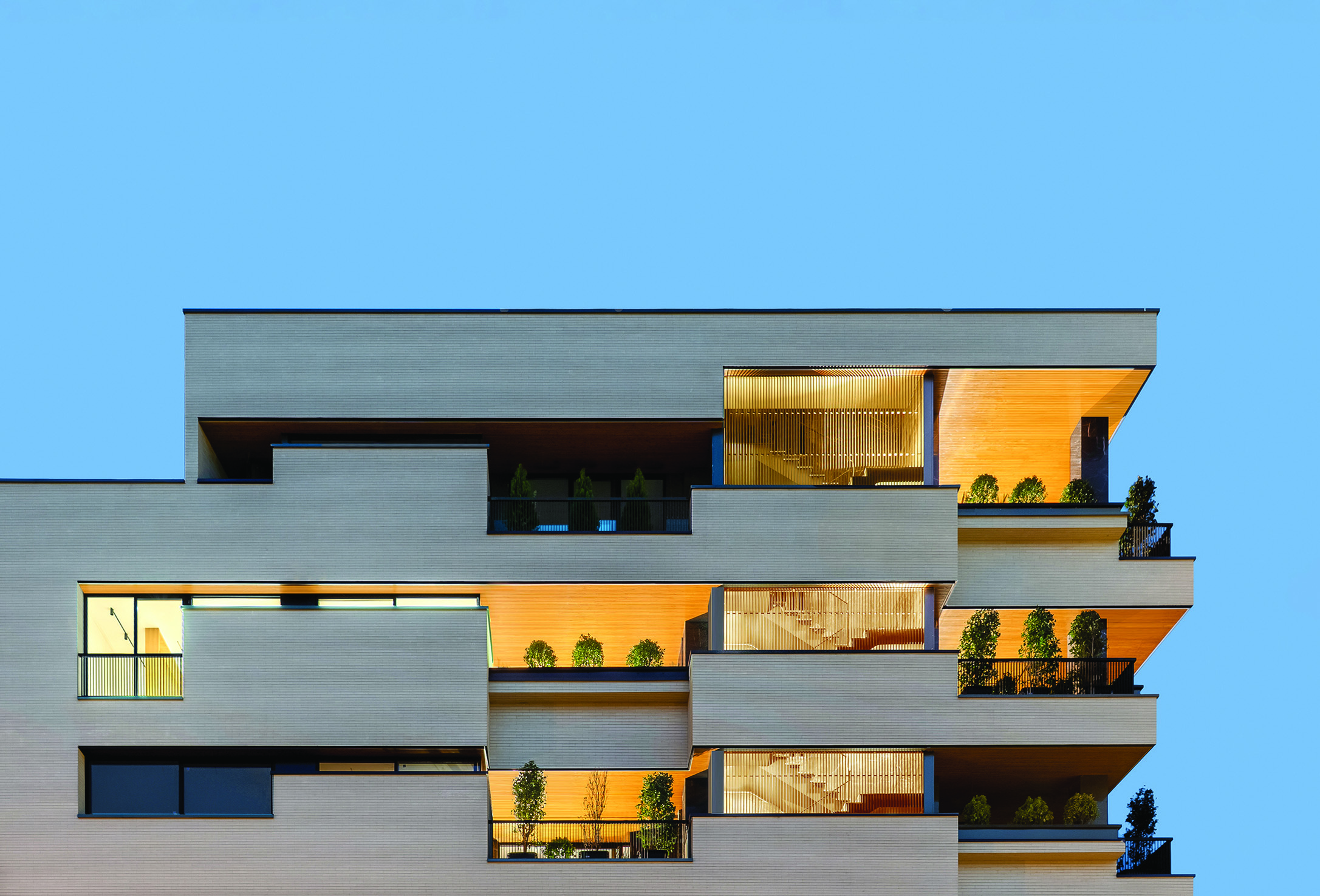 Full corner view of Bagh Mashhad at dusk, the multi-storey apartment building with staggered balconies illuminated in warm light, against a deep blue sky, neighbouring buildings visible