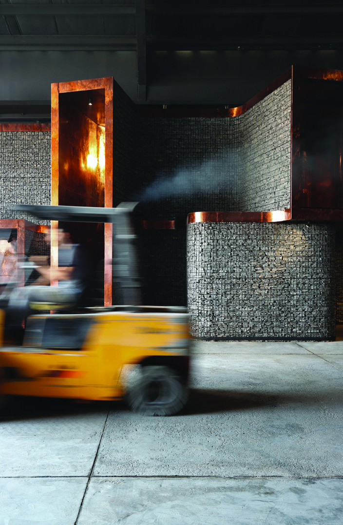 A yellow forklift in motion passes the gabion wall with its copper modules, the industrial rhythm of the factory continuing around the architecture
