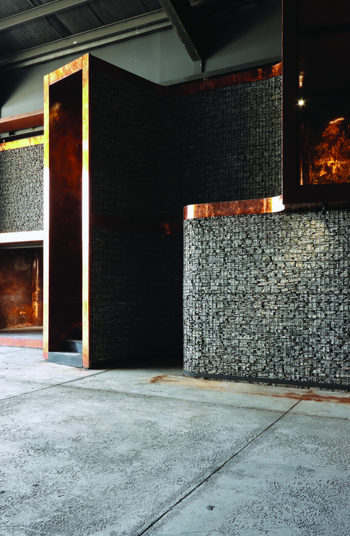 Close-up of the gabion wall and a copper-clad module &mdash; recycled stone waste stacked within wire mesh, with a patinated copper box cantilevered forward