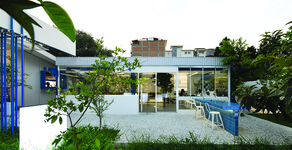 Garden view showing the renovated villa with white walls, metal roof, glass facades, blue-tiled outdoor bar counter with white stools, lush greenery, and gravel ground