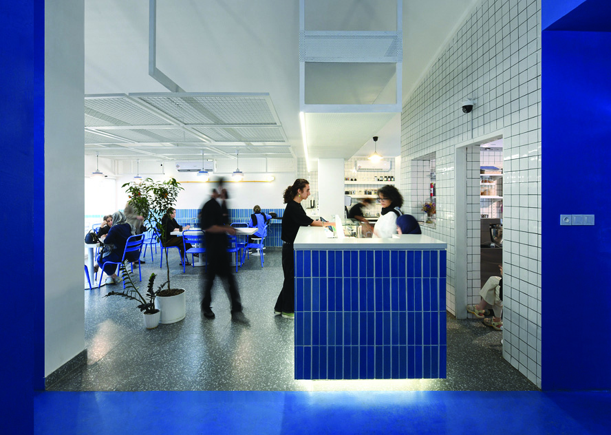 Interior view from the blue corridor looking into the main seating area with blue chairs and tables, bar counter, staff serving customers, plants, and mesh ceiling panels