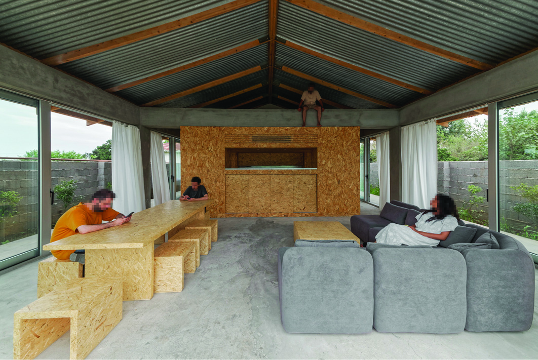 Wide interior view of Pati Project showing the central OSB-clad service box under the corrugated metal roof with exposed timber beams, grey sofas in the foreground, OSB dining table and stools, people relaxing