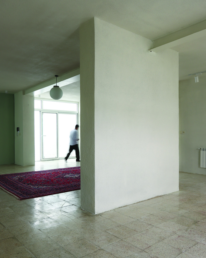 Interior view showing a thick white load-bearing wall dividing two rooms, a red Persian rug on the floor, a blurred figure walking past the glass courtyard door