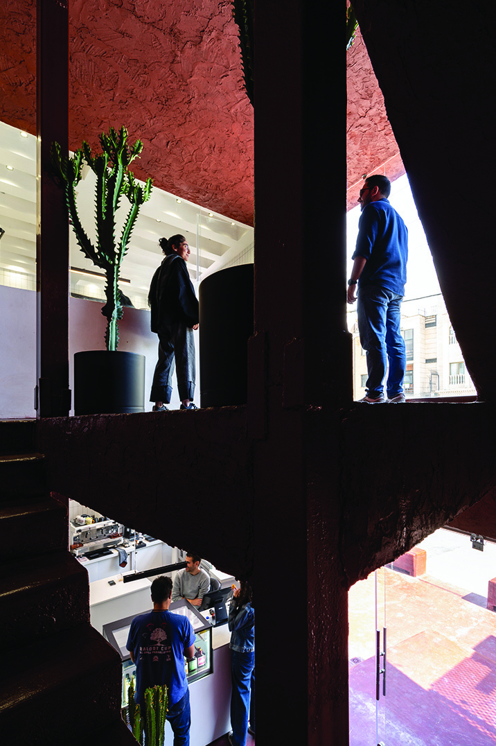 Split-level interior view of Journey Cafe: upper level shows two people standing in angular openings with red cement walls, lower level shows the bar counter with baristas at work
