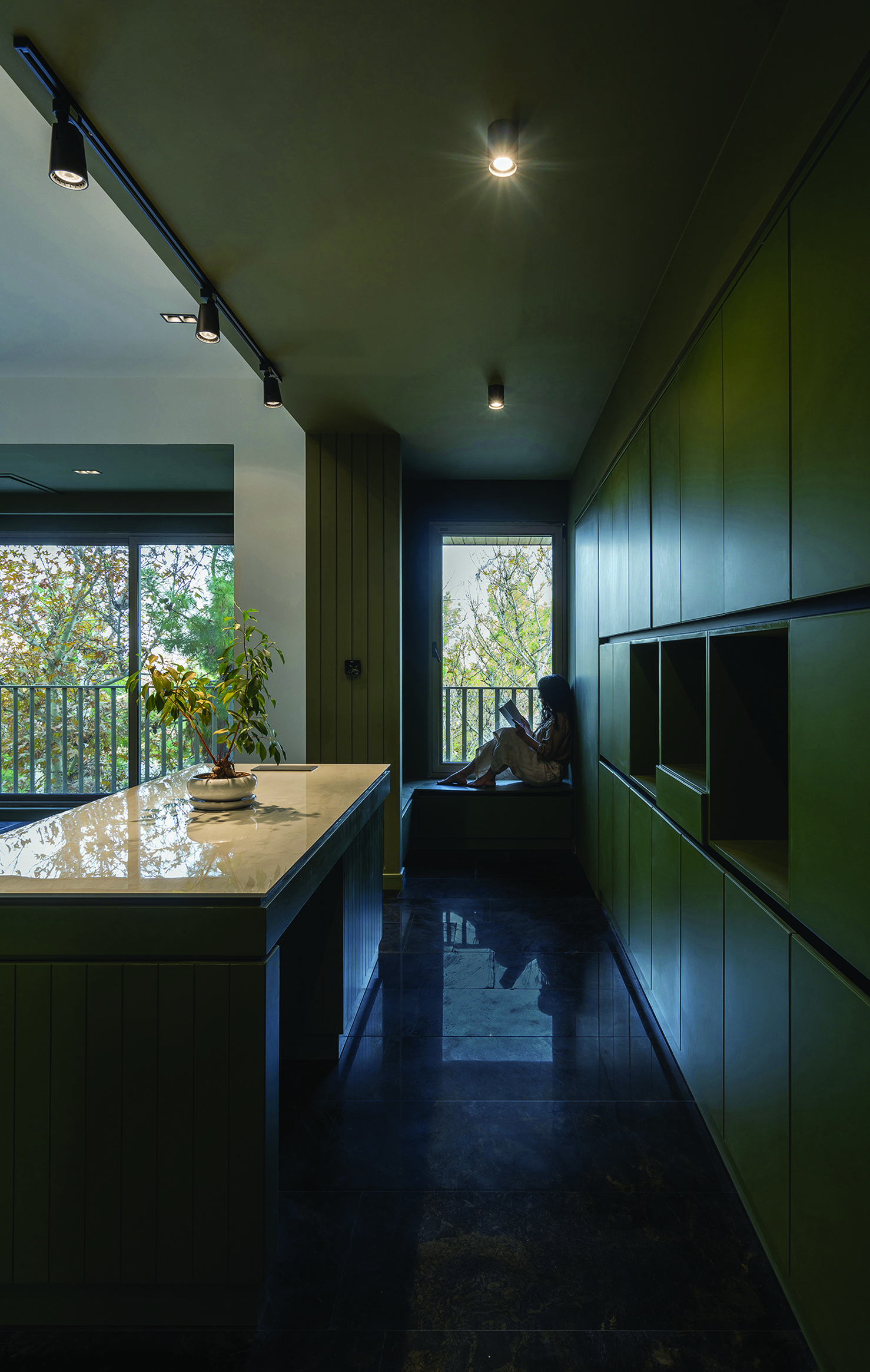 Dark green kitchen corridor with counter, track lighting, a person sitting in a window nook reading, trees visible through the window