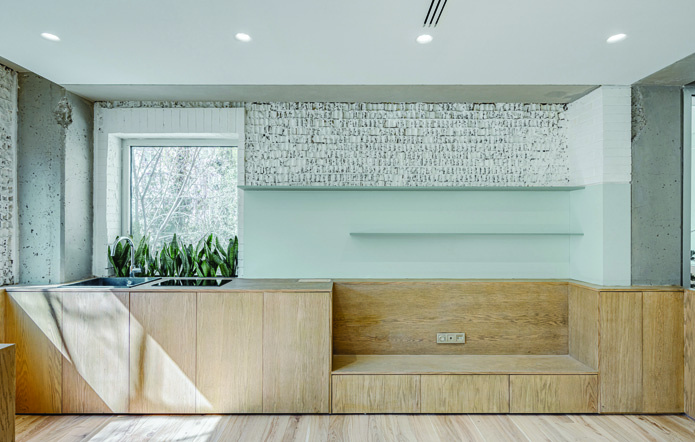 Kitchen with oak cabinetry, exposed whitewashed brick wall, concrete columns and beams, a window looking out to the garden with plants on the sill