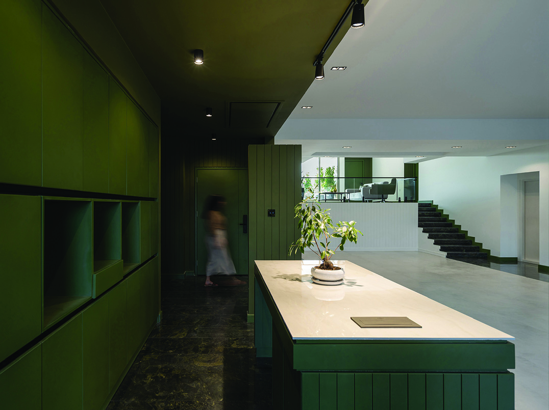 Green kitchen island with white stone counter and potted plant, looking toward the living room with staircase and glass railings visible beyond