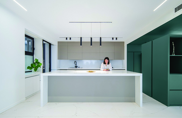 A minimalist kitchen with a white island counter, pendant lights, and a dark green cabinetry wall, a woman seated at the island, a potted plant by the corner window
