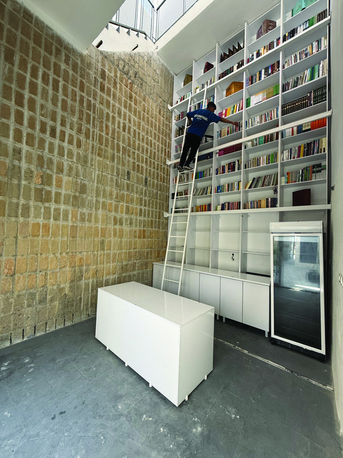 The library interior with a floor-to-ceiling white bookshelf filled with books, a person climbing a rolling ladder, a rough-hewn stone wall on the left, and a white central table below