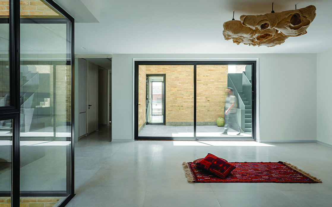 Interior of the living room with a red Persian rug on polished floor, sculptural organic light fixture, large glass sliding door opening to the brick courtyard with the green staircase visible
