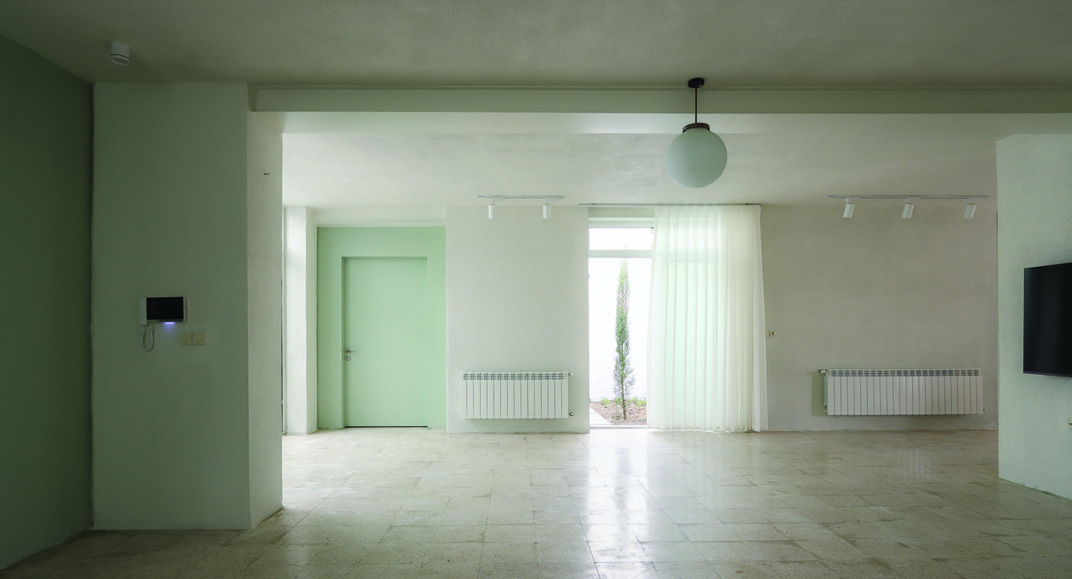 Ground-floor living room from the opposite angle: white walls with green door visible centrally, sheer curtains filtering daylight, a TV mounted on the right wall