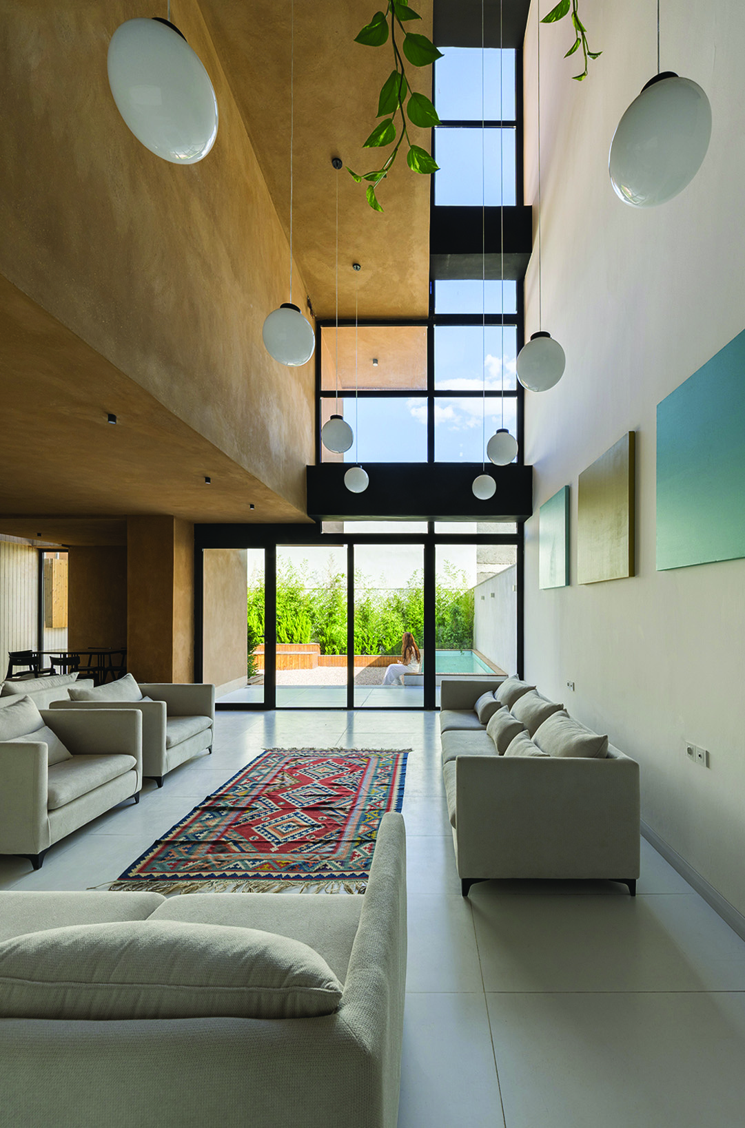 Double-height living room interior with earth-toned walls, pendant globe lights, a traditional Persian rug, sofas, and a full-height glass wall looking out to the pool and greenery