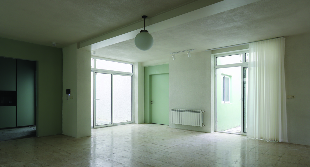 Interior of the ground-floor living room showing white textured walls, a green door to one side, pendant globe light, and full-height glass doors opening onto the courtyard