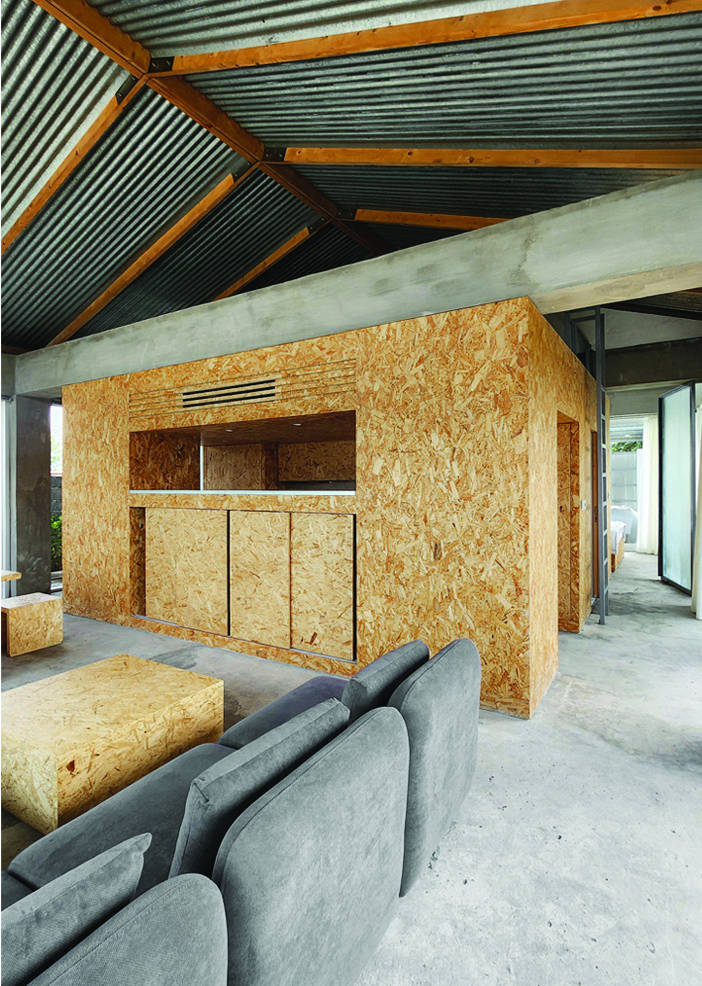 Interior view showing the OSB-clad service core with a pass-through window, corrugated metal ceiling with timber beams above, grey sofa in the foreground