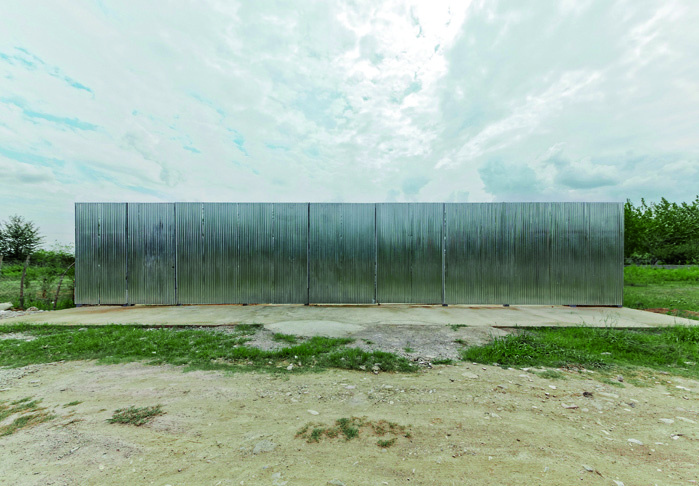 Corrugated metal entrance gate of Pati Project fully closed, forming a long silver wall on the concrete platform with green fields beyond